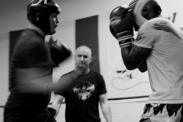 Zwei Boxer in Helmen und Handschuhen kämpfen in einem Ring, mit einem Schiedsrichter zwischen ihnen und einem Banner an der Wand im Hintergrund.