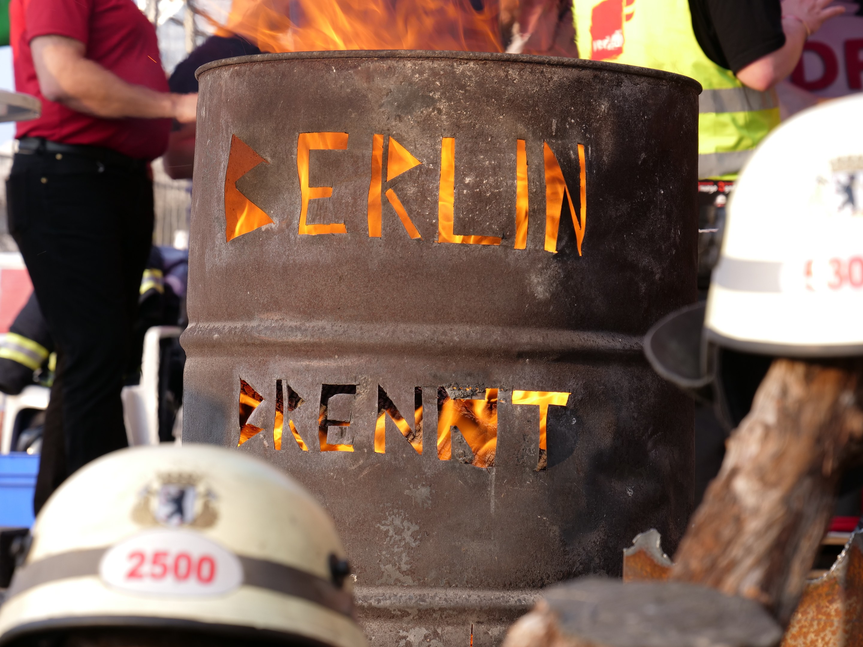 Eine Gruppe von Menschen steht um einen Feuerhydranten mit der Aufschrift "Berlin", mit einem brennenden Eimer im Vordergrund, Helmen unten und einem hölzernen Objekt auf der rechten Seite.