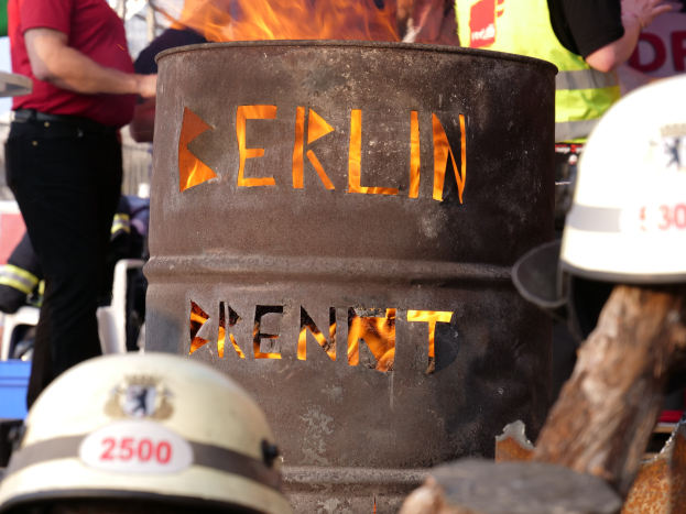 Eine Gruppe von Menschen steht um einen Feuerhydranten mit der Aufschrift "Berlin", mit einem brennenden Eimer im Vordergrund, Helmen unten und einem hölzernen Objekt auf der rechten Seite.