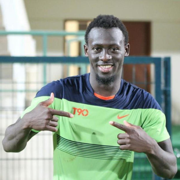 Ein grinsender Mann in einem grünen und blauen Hemd zeigt auf die Kamera, während ein Geländer, eine Wand und eine Tür leicht unscharf im Hintergrund zu sehen sind.