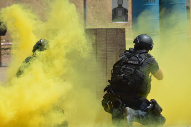 Zwei Polizisten in Einsatzausrüstung atmen gelben Rauch aus, während sie neben einer Wand mit Text auf einer Tafel und anderen Gegenständen im Hintergrund stehen.