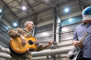Ein Mann in einer Militüruniform spielt eine Gitarre, whrend er neben einem anderen Mann in einem Helm steht, mit Rohren und Lichtern im Hintergrund.