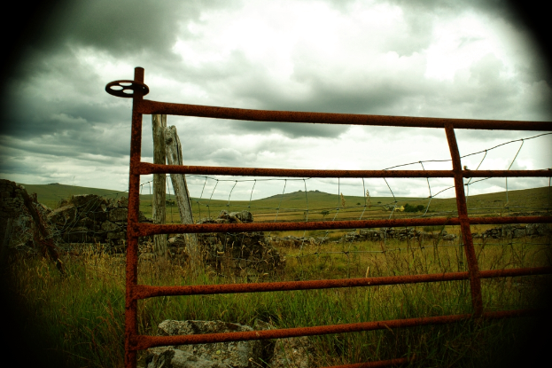 Eiserne Gitter im Vordergrund mit Holzpfählen, einem Zaun, Hügeln, Gras und einem bewölkten Himmel im Hintergrund.