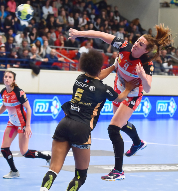 Frauen spielen Handball auf einem Feld mit einem Ball in der Luft, beobachtet von Zuschauern in einer Stadionatmosphäre.