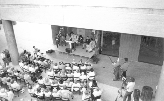 Ein Schwarz-Weiß-Bild eines Mannes, der auf einem Podium spricht, mit vielen Menschen, die auf Stühlen sitzen und zuhören, Fahnen im Hintergrund, einer Säule auf der linken Seite und Mülltonnen und einem Kameraständiger auf der rechten Seite.