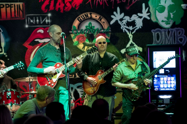 Drei Männer, die auf einer Bühne Gitarren spielen, mit einer Menge darunter und einer gemalten Wand im Hintergrund.