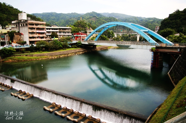 Eine Brücke spannt einen Fluss mit Gebäuden, Bäumen, Boden, Hügeln, Zäunen und Himmel im Hintergrund.