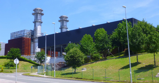 Ein großes Gebäude mit zahlreichen Fenstern, als Kraftwerk durch Beschriftung identifiziert, umgeben von Straßeninfrastruktur, Fahrzeugen, Vegetation und einem bewölktem Himmel.