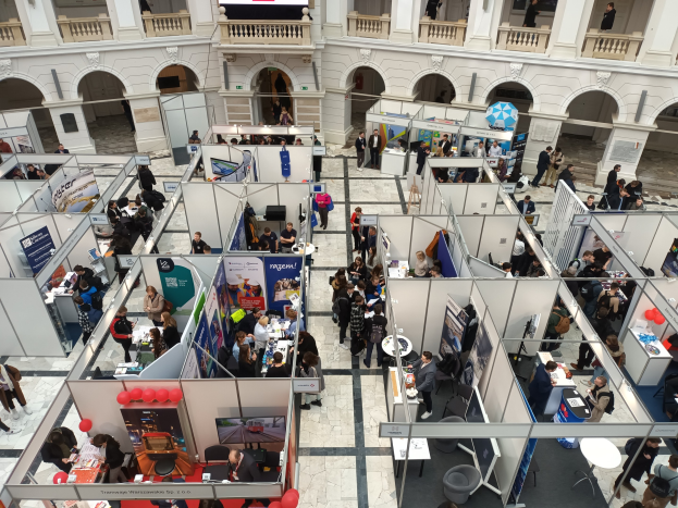 Eine große Gruppe von Menschen auf einer internationalen Messe mit Ständen, Bannern, Tischen, Stühlen, Luftballons und einem Gebäude mit Bögen, Säulen, Geländern und einem Bildschirm im Hintergrund.