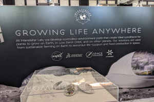 Ein Stand mit einem "Leben Wachsen lassen"-Schild, das einen Tisch mit Gegenständen im Vordergrund und eine Tafel mit Text und Bildern, Lampen und anderen Gegenständen im Hintergrund zeigt, Teil der Interstellar Lab-Ausstellung.