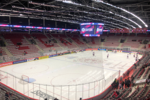 Ein Hockey-Spiel in einer großen Arena mit Zuschauern auf den Sitzplätzen und Stehplätzen, einem Zaun um die Eisfläche, einem Bildschirm mit der Aufschrift "St. Louis Blues vs Detroit Red Wings", Metallrahmen und einer Decke mit Deckenleuchten.