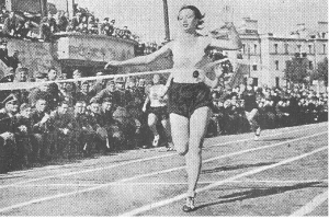 Ein Schwarz-Weiß-Foto einer Frau, die auf einer Laufbahn während des Frauen-100m-Finals bei den Olympischen Spielen 1956 läuft, mit einer Menschenmenge, Gebäuden und Pfosten im Hintergrund.