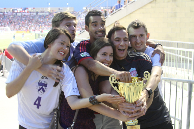 Sechs Menschen, darunter zwei Frauen und vier Männer, halten einen Pokal und lächeln für ein Foto vor einem Eisengeländer, mit einer Wand und einem weiteren Geländer oben rechts und Menschen und Lichtern im Hintergrund, möglicherweise in einem Stadion.