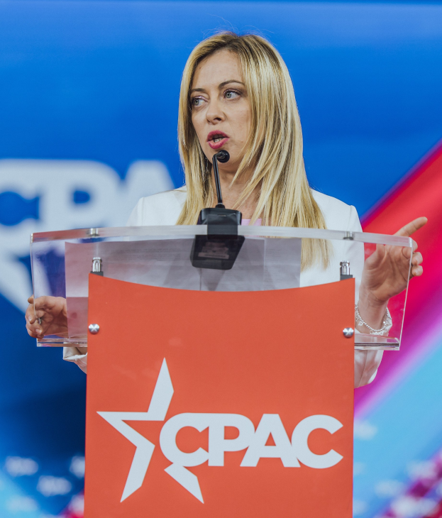 Frau in weißem Kleid, die an einem Rednerpult mit einem Mikrofon spricht, unscharfer Hintergrund, möglicherweise eine Politologin.