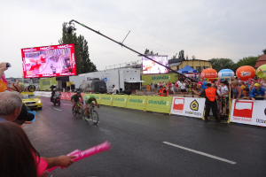 Eine Gruppe von Radfahrern fährt eine Straße entlang während der Tour de France, mit Zuschauern auf beiden Seiten, einer Kamera auf der linken Seite und Gebäuden, Bäumen und einem klaren blauen Himmel im Hintergrund.