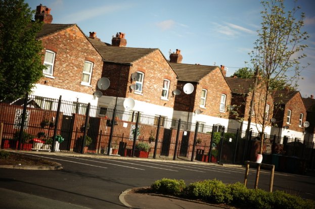 Gebäude mit Fenstern, Bäumen, einem Geländer, Blumenkübeln, einer Straße, dem Himmel und Satellitenschüsseln an der Wand befestigt.