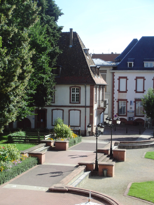 Ein Rathaus mit einem zentralen Brunnen in einem Park, umgeben von Geb├Ąuden, Stra├čenm├Âbeln, Grünfl├Ąchen und einem bew├Âlktem Himmel.