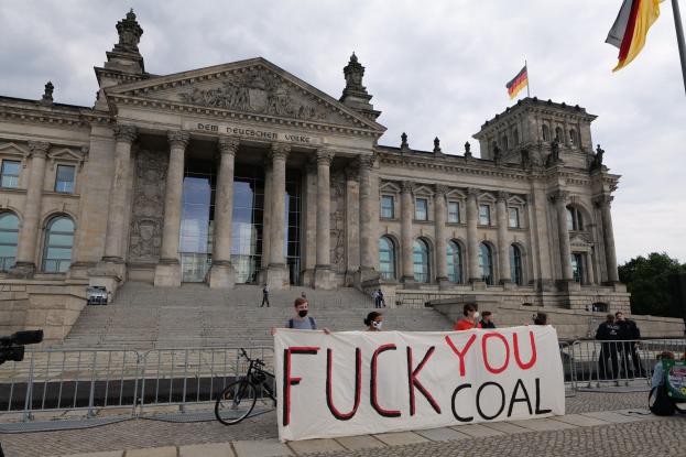 Gruppe von Menschen mit einem "Fuck You Coal"-Schild vor dem Reichstag in Berlin, mit Bäumen, Fahnenmast und bewölktem Himmel im Hintergrund.
