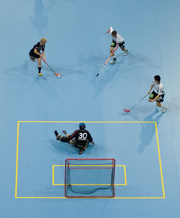 Drei Personen in Sportbekleidung spielen Hockey, wobei eine Person rechts den Ball mit einem Hockeystock schlägt und eine Person hinter einem Tor auf dem Boden sitzt.