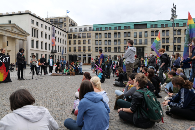 Eine Gruppe von Menschen, die auf dem Boden vor einer Menge mit Fahnen und Spruchbändern sitzen, während einer Anti-Schwulen-Demonstration in Berlin. Im Hintergrund sind eine Statue, Gebäude und ein bewölkter Himmel zu sehen.
