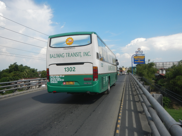 Ein grüner und weißer Bus fährt auf einer Autobahn mit Geländern, Bäumen, Gebäuden, Texttafeln, Polen, Drähten und einem klaren blauen Himmel im Hintergrund.