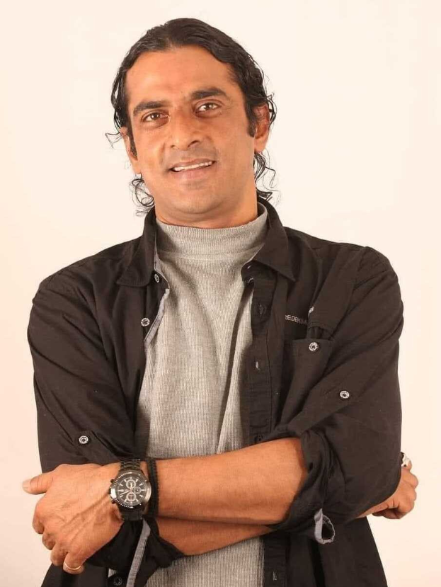 Ein Mann in einem schwarzen und aschfarbenen Kleid mit verschr├Ąckten Armen, der eine Uhr tr├Ągt, vor einem wei├čen Hintergrund steht.