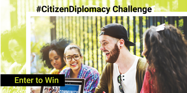 Eine Gruppe von Menschen versammelt sich im Freien um einen Laptop mit der Aufschrift "Citizen Diplomacy Challenge - Gewinnen Sie", mit einem Mann in der Mitte, der eine Mütze und eine Schutzbrille trägt und einen Stift hält, während die anderen lächeln und in ein Gespräch vertieft sind, mit Bäumen und einem Zaun im Hintergrund.