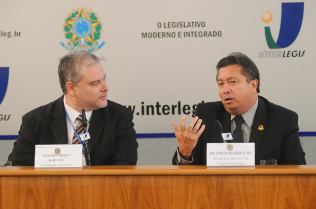 Zwei Männer in Anzügen sitzen an einem Tisch mit zwei Namensschildern und zwei Mikrofonen; Logos und Text sind im Hintergrund sichtbar.