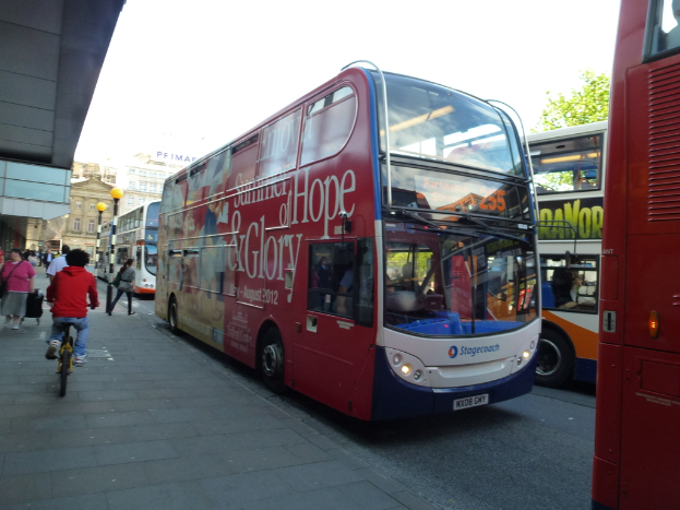 Eine Person fährt ein Fahrrad auf einem Gehweg mit vielen Menschen, Bussen mit Text darauf, Gebäuden und Laternen im Hintergrund.