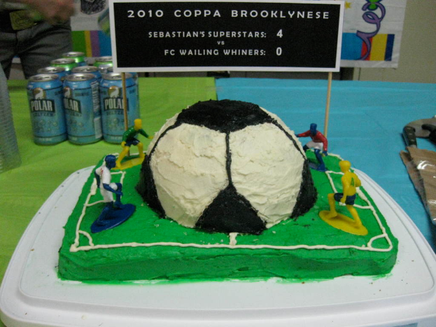 Ein Fußballstadion-Kuchen mit einem Fußball und vier Spielern, einer Fahne, Softdrink-Dosen und einer Person in der oberen linken Ecke.