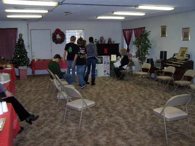 Ein Raum mit mehreren Stühlen, Tischen und Menschen, einige stehen, einige sitzen, mit Tassen und Gefäßen auf den Tischen, einem Weihnachtsbaum, einem Piano auf der rechten Seite, Foto Rahmen, Fenstern und Röhrenleuchten an der Decke und einer Wand im Hintergrund mit einem Bouquet, Vorhang, Tür und weiteren Foto Rahmen.