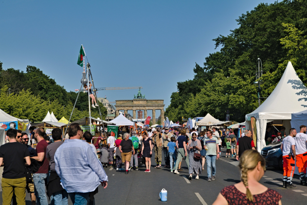 Menschenmenge auf einer Straße mit Zelten, Fahrzeugen und Bäumen während eines Events mit Fahnen, einem Bogen im Hintergrund und einem klaren blauen Himmel.
