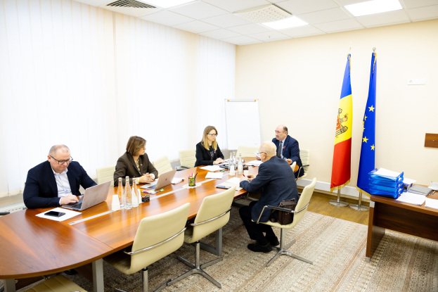 Eine Gruppe von Menschen sitzt um einen Konferenztisch mit Laptops, Flaschen, Gläsern, Papieren, Büchern und anderen Gegenständen, die in eine Diskussion vertieft sind, mit Flaggen, einer Whiteboard und Deckenleuchten im Hintergrund.