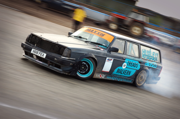 Ein Auto rast auf einer Rennstrecke, wobei Rauch ausstie├čt, mit anderen Fahrzeugen und Menschen im Hintergrund.