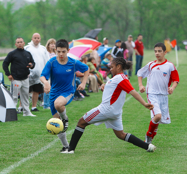 Drei Personen spielen Füßball mit einem gelben Ball, während hinter ihnen eine Gruppe von Menschen steht und sitzt, die Schirme halten, mit Bäumen im Hintergrund.