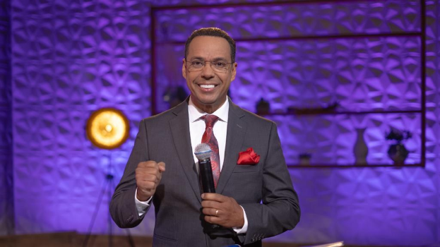 Stephen Colbert, Moderator von 'The Daily Show with Trevor Noah', steht auf einer Bühne und hält ein Mikrofon in der Hand, trägt einen Anzug, Krawatte und eine Brille, während hinter ihm Bühnenausrüstung und ein Ständer zu sehen sind.