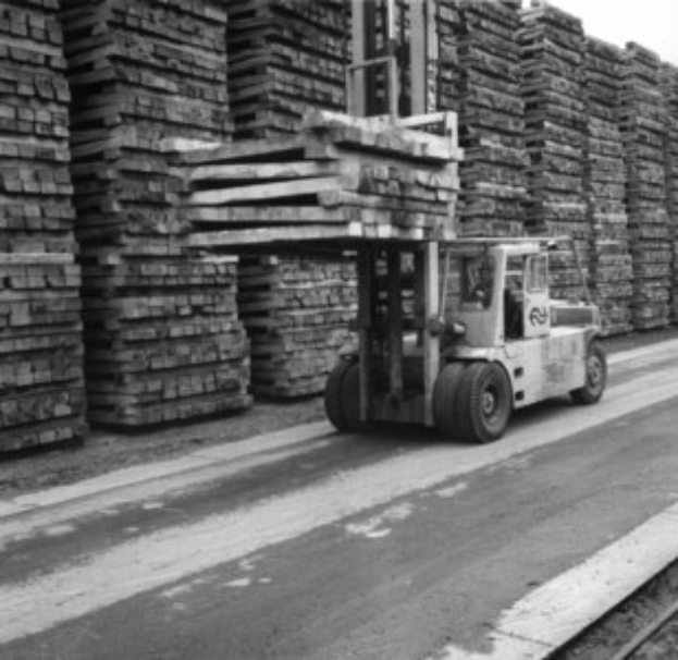 Schwarz-weißes Foto eines Gabelstaplers mit Fahrer auf einer Straße neben Bahnschienen, mit gestapelten Holzplanken im Hintergrund.