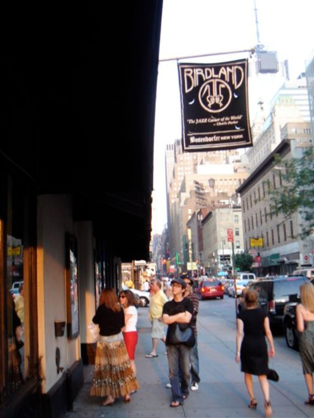 Eine belebte Straßenszene mit zahlreichen Fahrzeugen, Gebäuden auf beiden Seiten, einigen Fußgängern, die die Straße entlanggehen, und einigen Menschen, die vor einem Geschäft auf der linken Seite stehen.