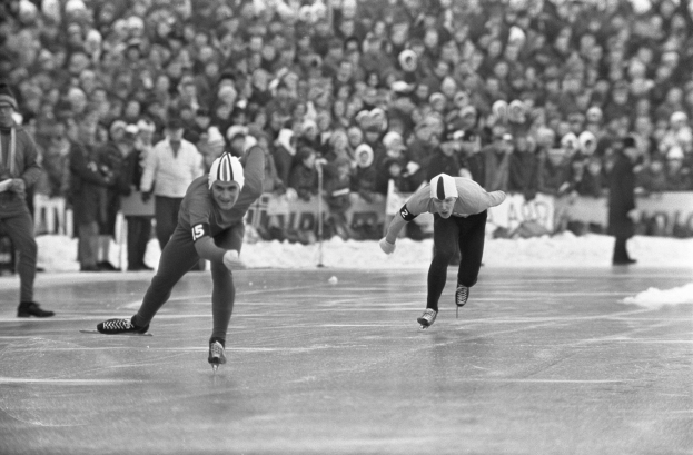 Zwei Männer im Eisschnelllauf auf einer Eisbahn vor einem großen Publikum, die Helme und Eislaufschuhe tragen, mit einem unscharfen Hintergrund, in Schwarz-Weiß.