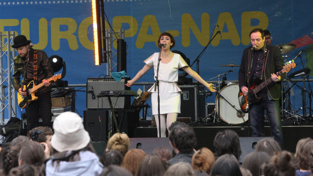 Eine Bühne mit vier Musikern: eine Frau in einem weißen Kleid singt in der Mitte, zwei Männer spielen Gitarren auf der linken und rechten Seite, eine Menschenmenge vorne und Lautsprecher im Hintergrund.