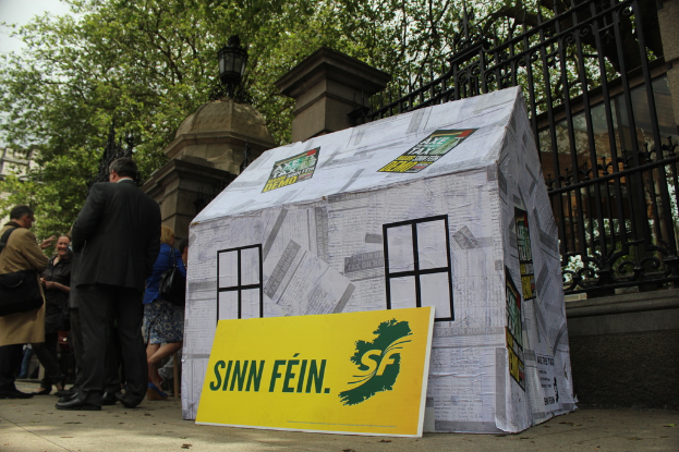 Ein Zelt mit einem Werbeplakat davor, umgeben von einer Menge, mit einem Eisengitter, Säulen, Straßenlaternen, Bäumen und Himmel im Hintergrund.