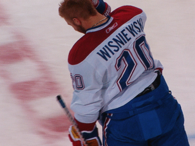 Ein Eishockeyspieler in einem "Wisniewski"-Trikot steht auf dem Eis, mit einem Hockeystock in der Hand und einem konzentrierten und entschlossenen Gesichtsausdruck.