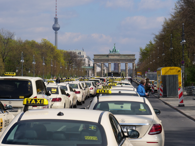 Eine belebte Straße in Berlin, Deutschland, gesäumt von parkenden Taxis, Fußgängern auf dem Gehweg, Laternen, Bäumen und Gebäuden, mit einem fernen Bogen mit Statuen und einem Turm unter einem bewölkten Himmel.