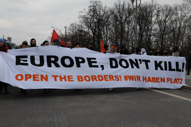 Eine Gruppe von Menschen marschiert mit einem Banner, auf dem "Europa, töte nicht, öffne die Grenzen" steht, durch die Straße, während einige Flaggen schwenken, unter einem klaren blauen Himmel mit Bäumen und Laternen im Hintergrund.