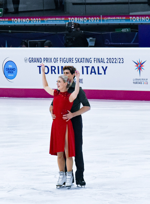 Zwei Eiskunstläufer, Tessa Virtue und Scott Moir, zeigen eine Figur auf einem Eisstadion, wobei Virtue ein rotes Kleid trägt und Moir ein schwarzes Hemd und Hosen, während Richter und Zuschauer im Hintergrund beobachten.