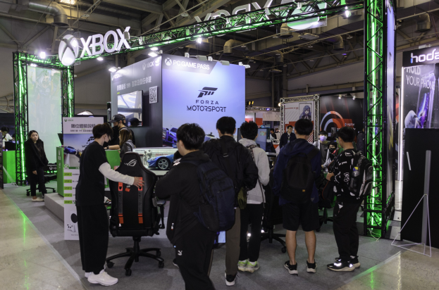 Eine Gruppe von Menschen steht vor einem Messestand, der "Forza Motorsport 7" mit Stühlen, Plakaten, Lichtern und anderen Gegenständen drumherum zeigt.