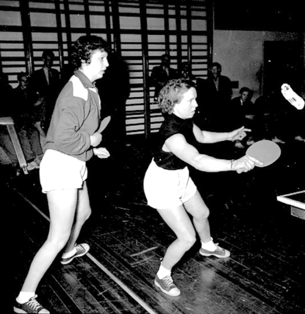 Zwei Frauen beim Tischtennis auf einem Court, jede hält einen Schläger in der Hand, mit einigen Zuschauern im Hintergrund und einem Tischtennis-Tisch auf der rechten Seite, dargestellt in Schwarz-Weiß.