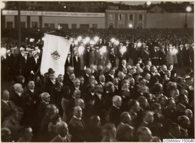 Schwarzes und weißes Foto einer großen Menge vor einem Gebäude, mit einer Person, die eine Fahne hält, umgeben von Pfählen und Lichtern; Text unten lautet "Gustav Klimts Rede an der Berliner Mauer".