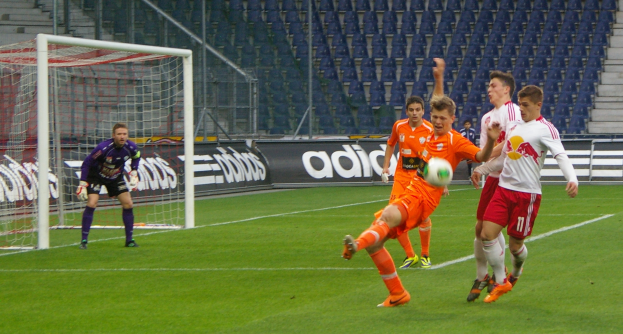 Eine Gruppe von Männern, die auf einem Feld Fußball spielen, mit einem Torpfosten auf der linken Seite, Bannern mit Text im Hintergrund und sichtbaren Stühlen und Treppen.