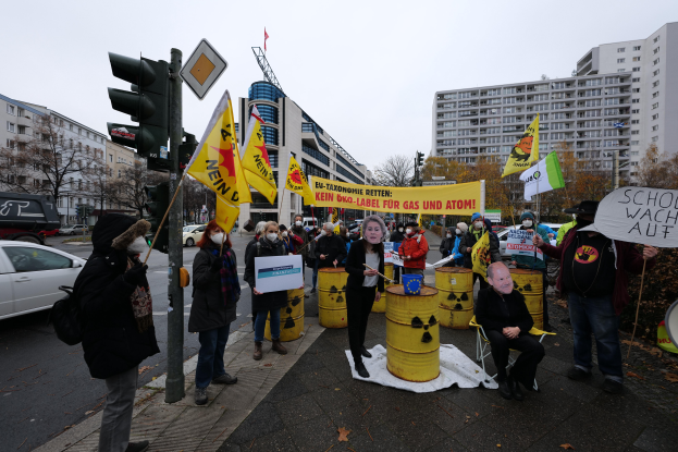 Eine Gruppe von Menschen steht auf einer Straße und hält gelbe Fässer und Schilder hoch, mit einer Person im Vordergrund und Fahrzeugen, Verkehrszeichen, Schildern, Bäumen, Gebäuden und einem klaren blauen Himmel im Hintergrund, die scheinbar gegen Atommüll protestieren.